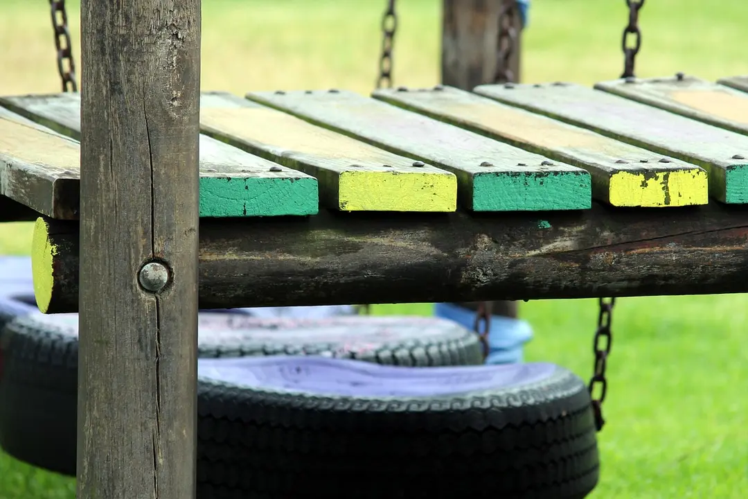 Close-up of a wooden playground platform with painted green and yellow slats, with hanging tires in a grassy park