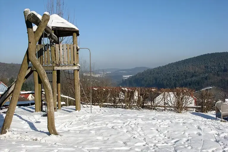 Snow-covered wooden playground structure in a residential area with distant hills and trees