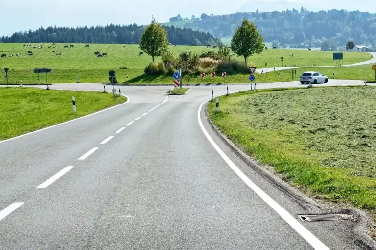 Two-lane rural road intersection with green fields, trees, and road signs in the distance.