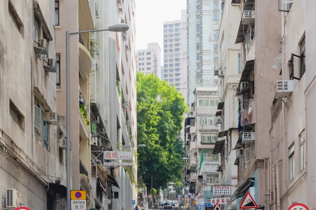 Urban city street between tall residential buildings, with parked cars and a tree-lined avenue in the distance.