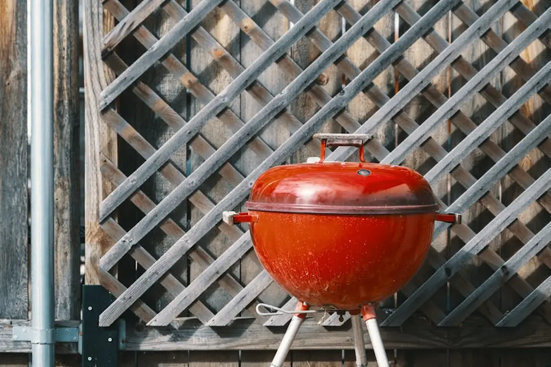 Red kettle-style outdoor grill on a stand in front of a wooden lattice fence.