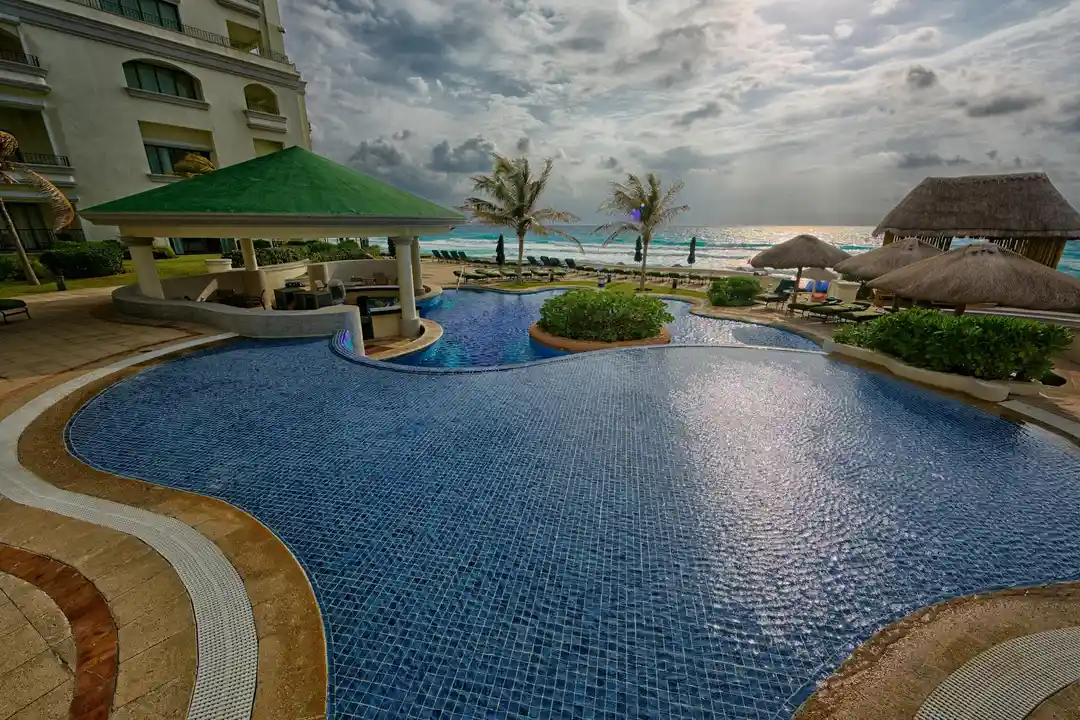 Curved resort pool with blue tiles, palm trees, cabanas, and a beach/ocean view in the background