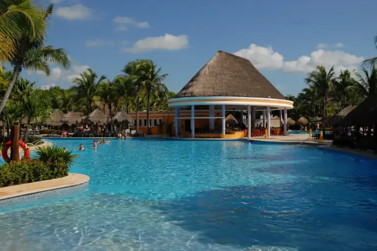 Resort-style outdoor pool with palm trees and a thatched-roof pool bar under a blue sky.