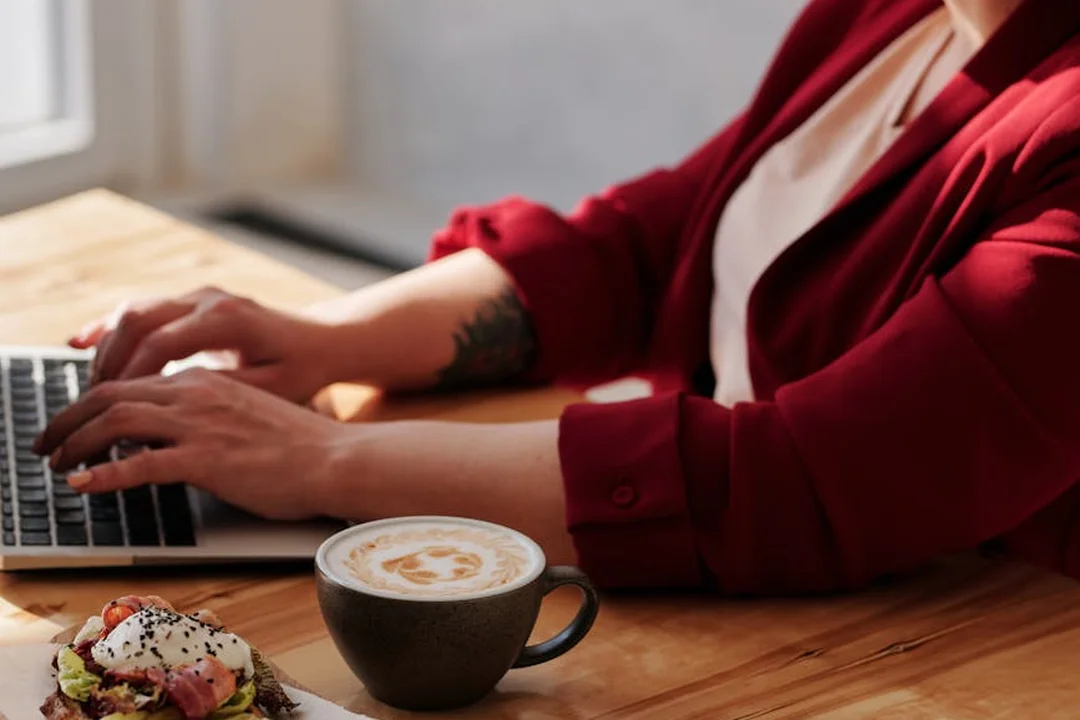 Person typing on a laptop at a wooden table with a cup of coffee and a pastry nearby.