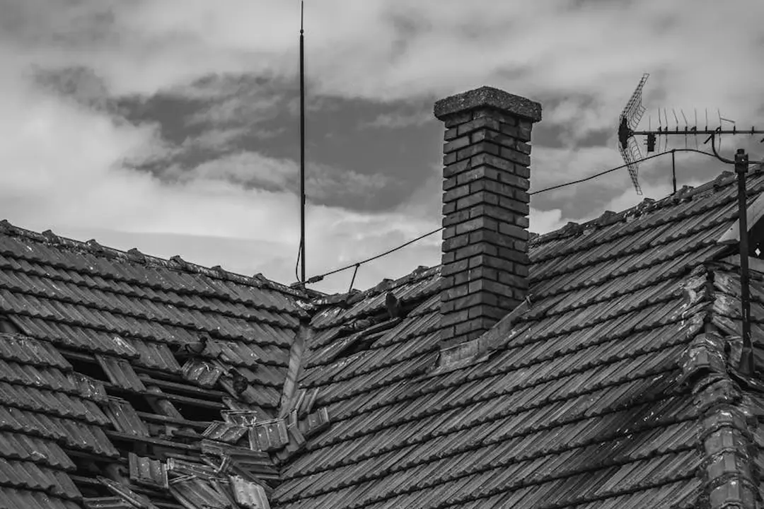Black-and-white photo of a deteriorating tiled roof with a brick chimney, an antenna, and a cloudy sky.