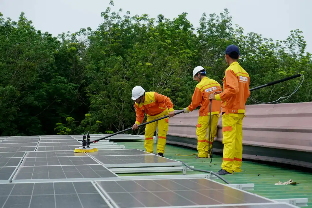 Three workers in bright orange and yellow safety outfits install solar panels on a building rooftop, with trees in the background.
