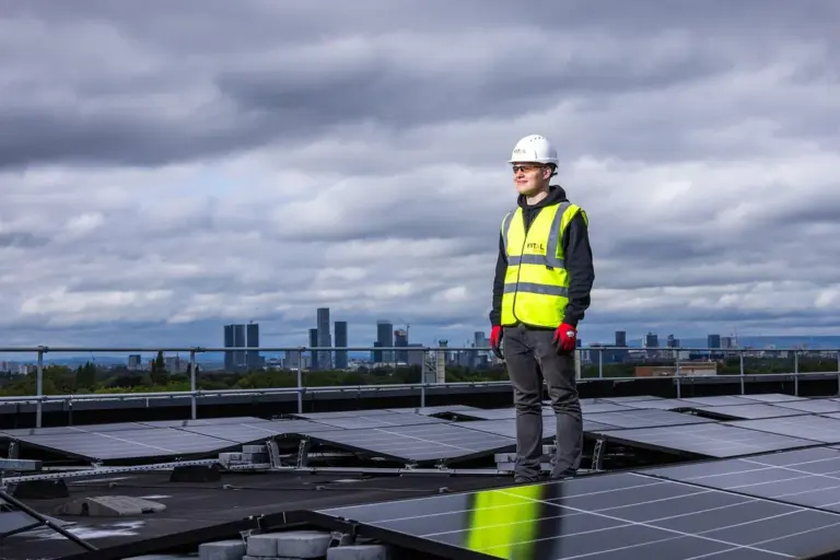 A safety worker stands on a rooftop among solar panels with a city skyline in the distance under a cloudy sky.