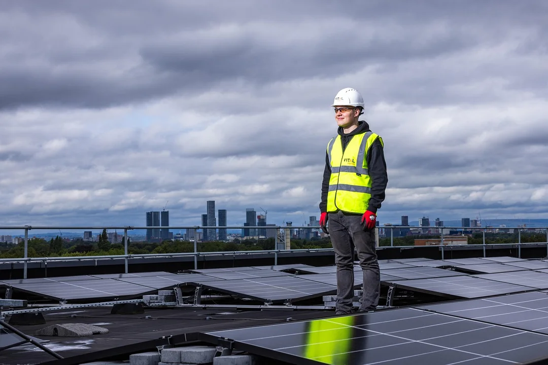 A safety worker stands on a rooftop among solar panels with a city skyline in the distance under a cloudy sky.