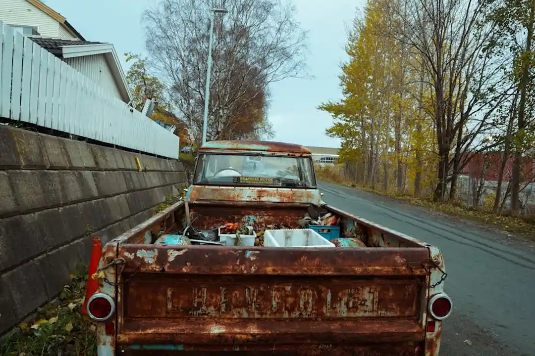 Rusty, abandoned pickup truck parked on a roadside with autumn trees in the background, illustrating vehicle abandonment and potential HOA towing scenarios.