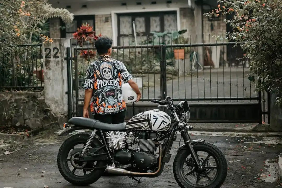A person stands beside a motorcycle in front of a gated residential property with a driveway and garden.