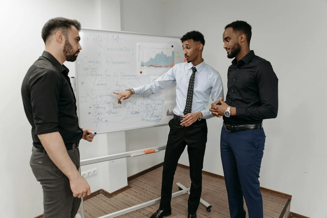 Three professionals in business attire standing around a whiteboard with charts in a conference room.