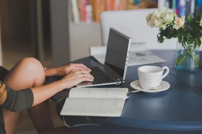 Person typing on a laptop at a desk with a notebook, cup of coffee, and flowers.