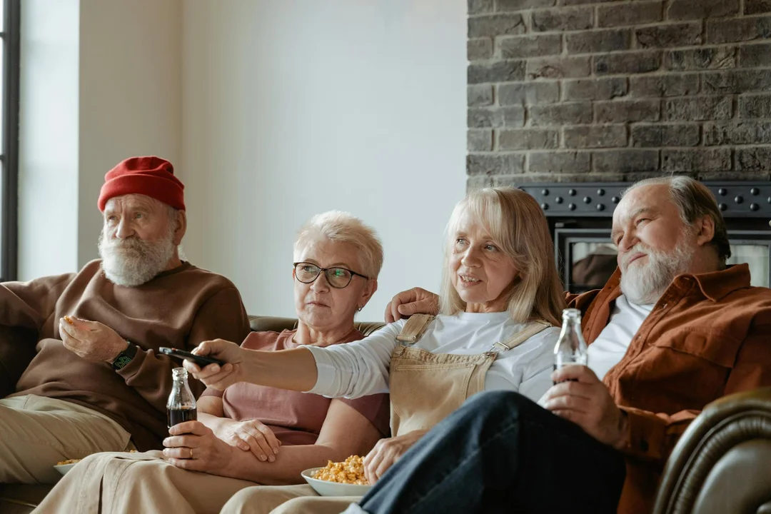 Four older adults sit on a sofa in a living room, with a remote control and drinks, illustrating a group-home common area.