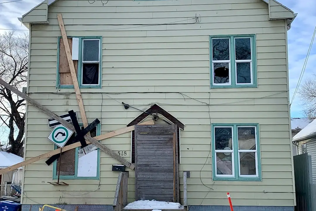 Two-story beige house with a boarded front entrance and broken windows, showing damage and debris around the doorway.