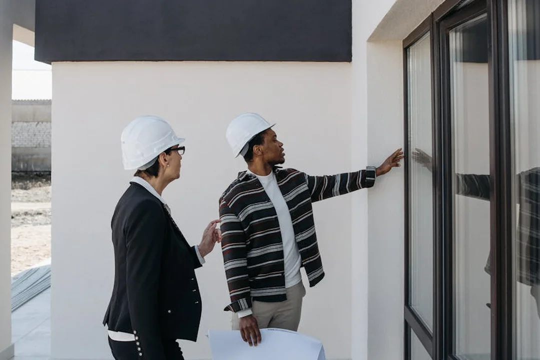 Two construction professionals wearing white hard hats review architectural plans near a building's glass door.