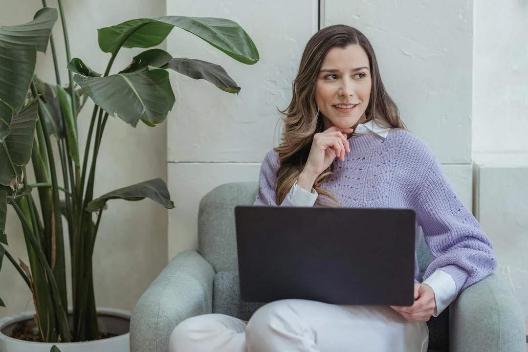 Woman sitting on a light gray sofa with a laptop, in a modern living room with a large plant in the background.