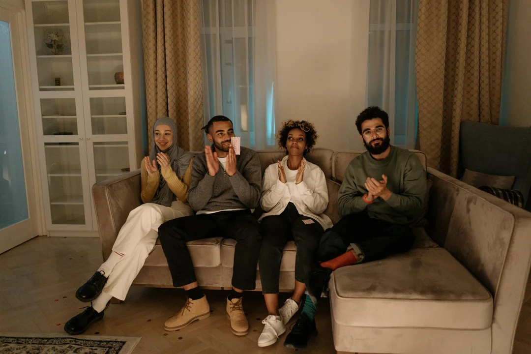Four adults seated on a living room couch, clapping and smiling, in a cozy home setting with curtains and shelves.