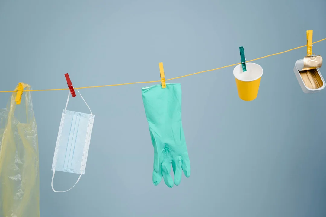 A clothesline against a soft blue background with a disposable face mask hanging by a red clothespin, a green glove hung by a green clip, a yellow cup clipped to the line, and a clothespin at the far right.