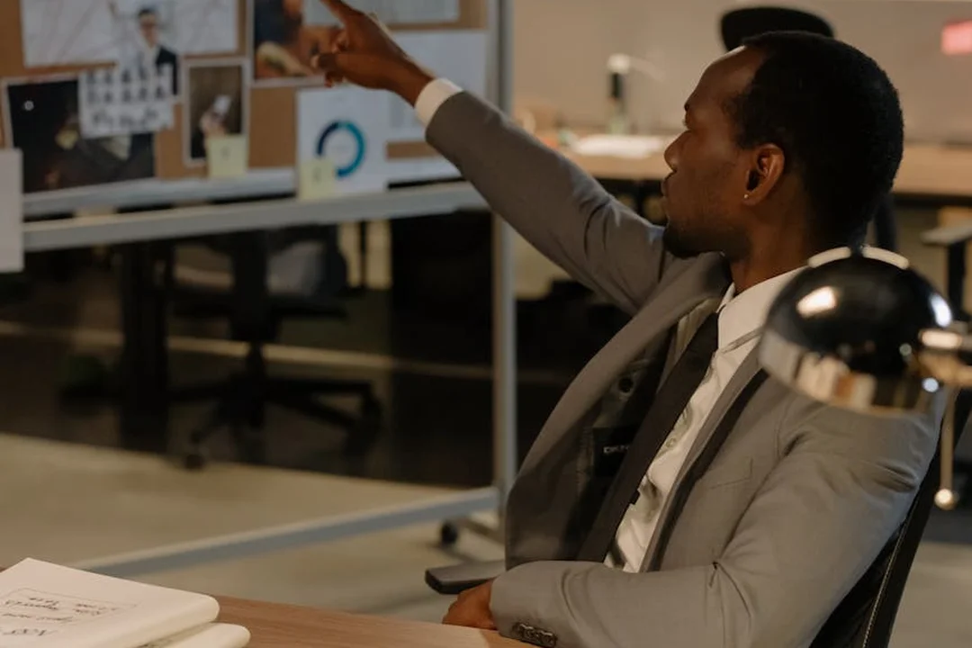 Business professional in a suit sits at a desk, pointing toward computer monitors displaying charts in an office.