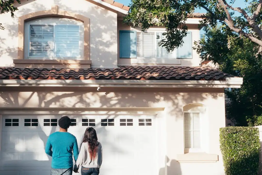 Front view of a two-story suburban house with a white garage door; a couple stands in the driveway facing the home.