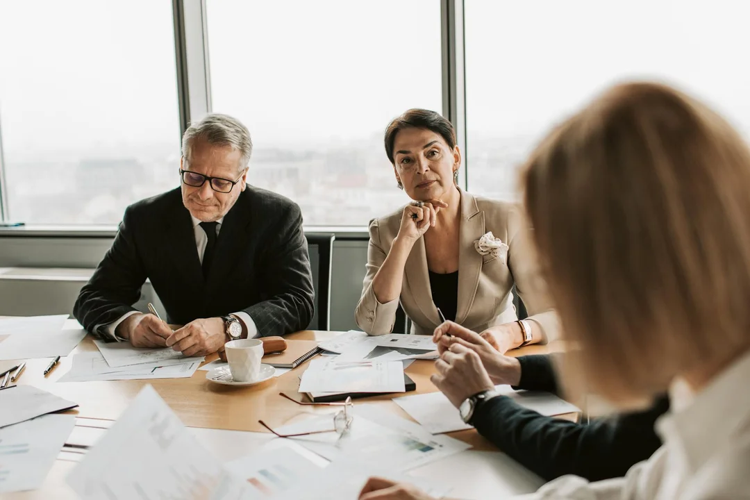 Three professionals sit at a conference table covered with documents; a fourth person in the foreground is out of focus, suggesting a tense meeting.