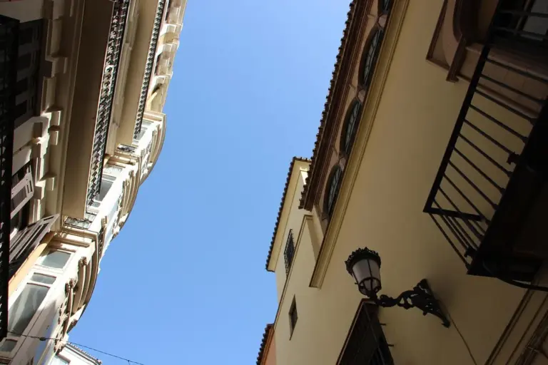 Upward view of a narrow street between tall residential buildings against a clear blue sky, with a decorative streetlamp on the right