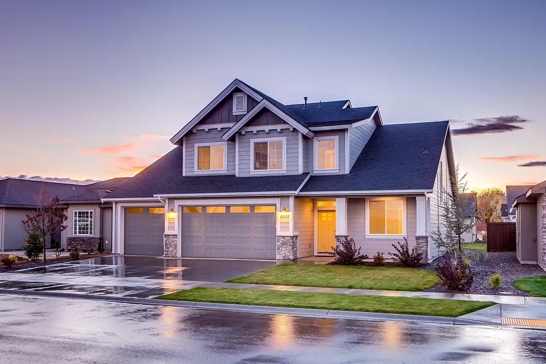 Two-story suburban home with a three-car garage and a well-kept lawn, photographed on a wet street at sunset.