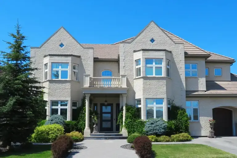 Front view of a large beige two-story house with manicured lawn, shrubs, and a small balcony, under a bright blue sky.