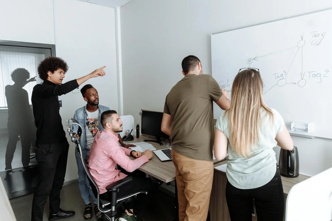 A diverse group of professionals gathered in a conference room, standing and seated around a table while reviewing notes on a whiteboard