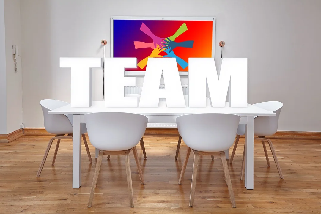 A modern conference room with a white table and white chairs arranged around it; a large TEAM sculpture sits on the table, and a colorful abstract painting hangs on the wall behind.