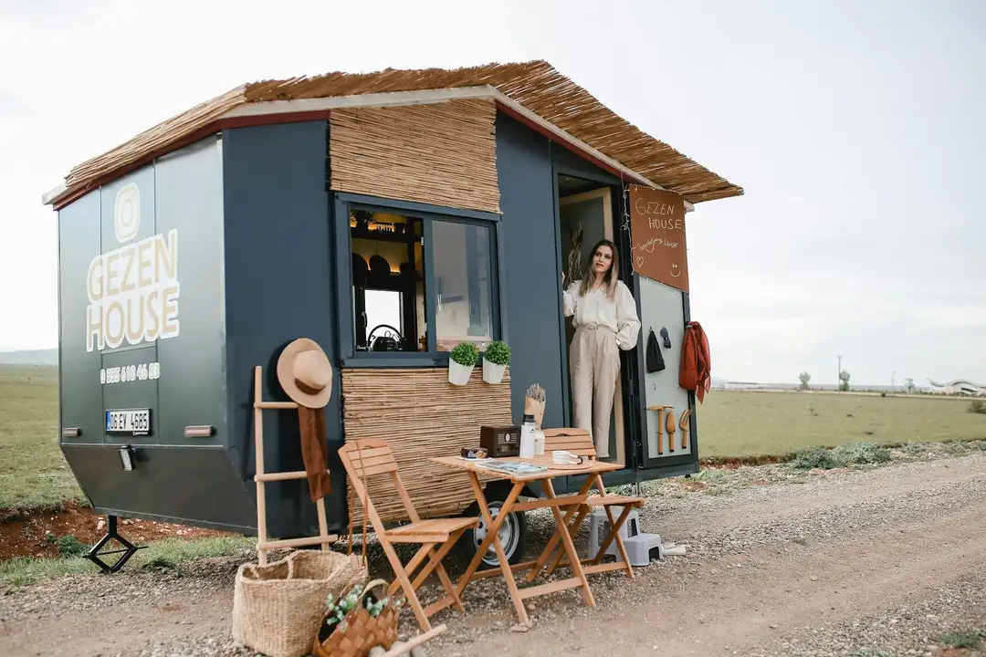 Blue tiny house on wheels with a small outdoor seating area and a person standing at the doorway in a rural landscape