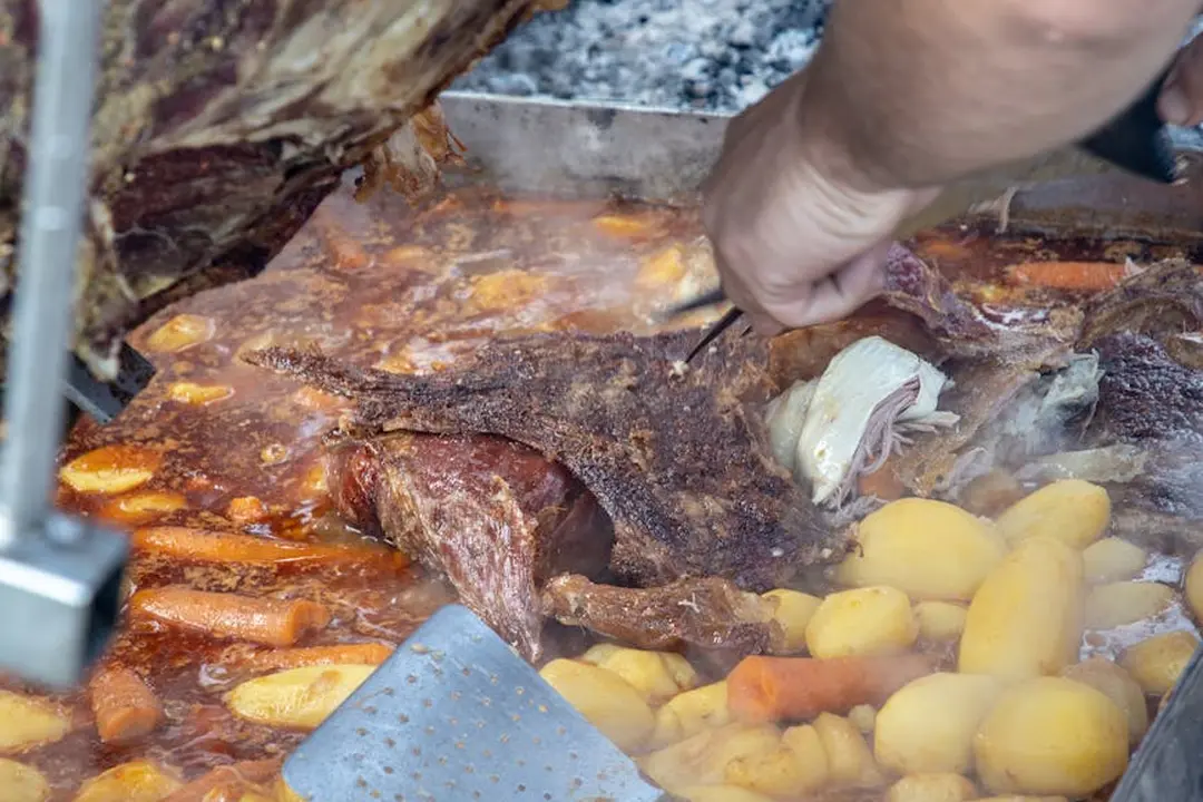 Close-up of a Traeger-style grill cooking ribs and corn outdoors, with a hand tending the food.