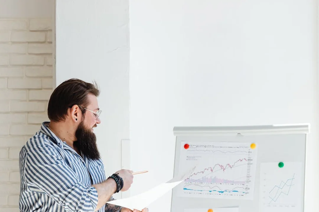 Bearded man in a striped shirt pointing at charts on a whiteboard in an office setting