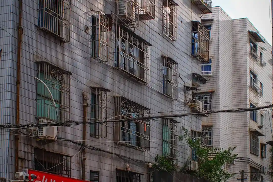 Multi-story urban apartment building with numerous windows, window air conditioning units, and metal security bars on some windows.