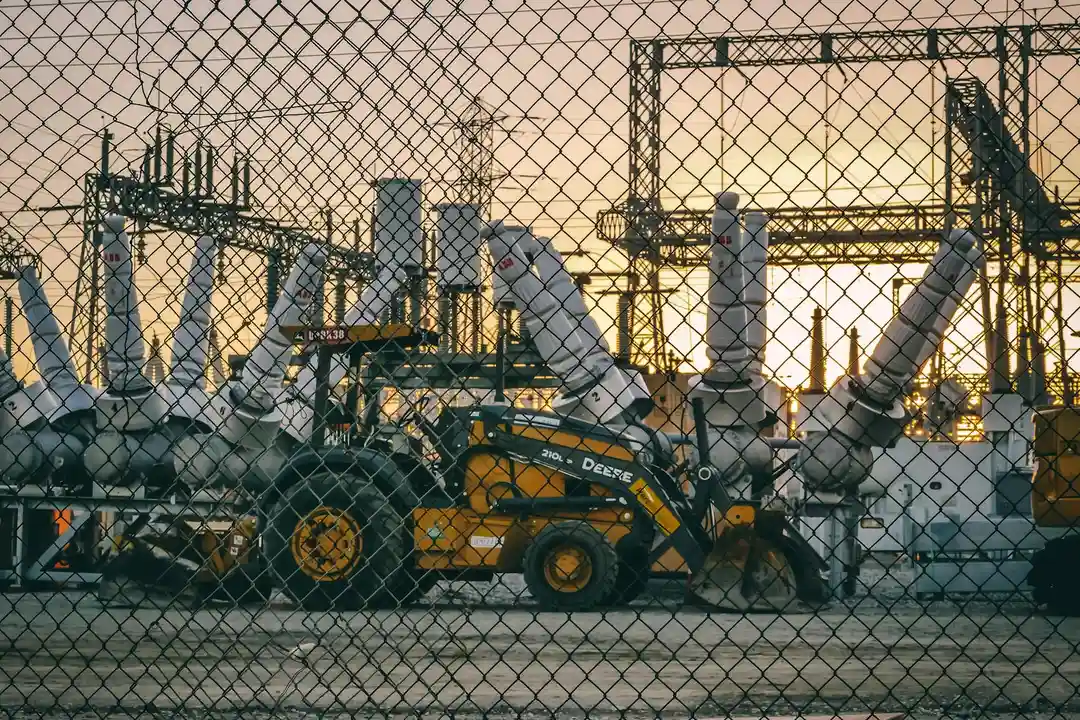 Industrial utility site behind a chain-link fence with heavy equipment and power infrastructure.
