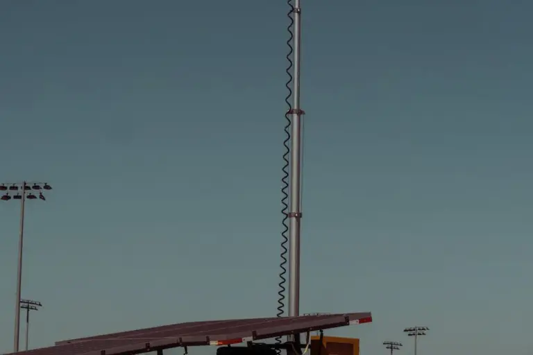 A tall vertical utility pole with a coiled cable against a clear blue sky, with a small carport-like structure in the foreground.