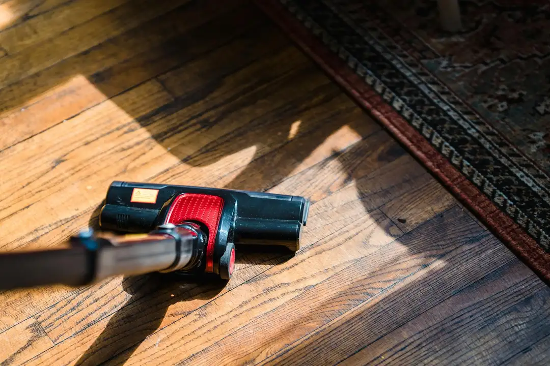 Cordless vacuum cleaner on a wooden floor beside a rug with sunlight casting shadows