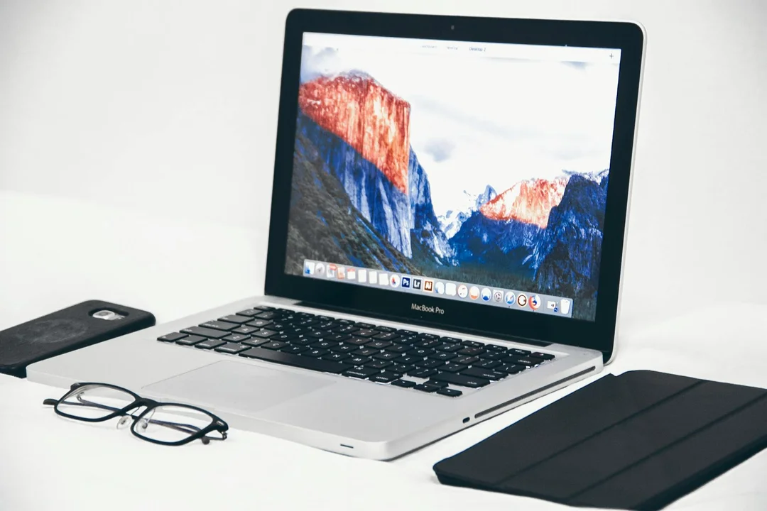 Laptop on a clean white desk with a pair of glasses and a closed black notebook, suggesting a workspace for managing vendors and maintenance.