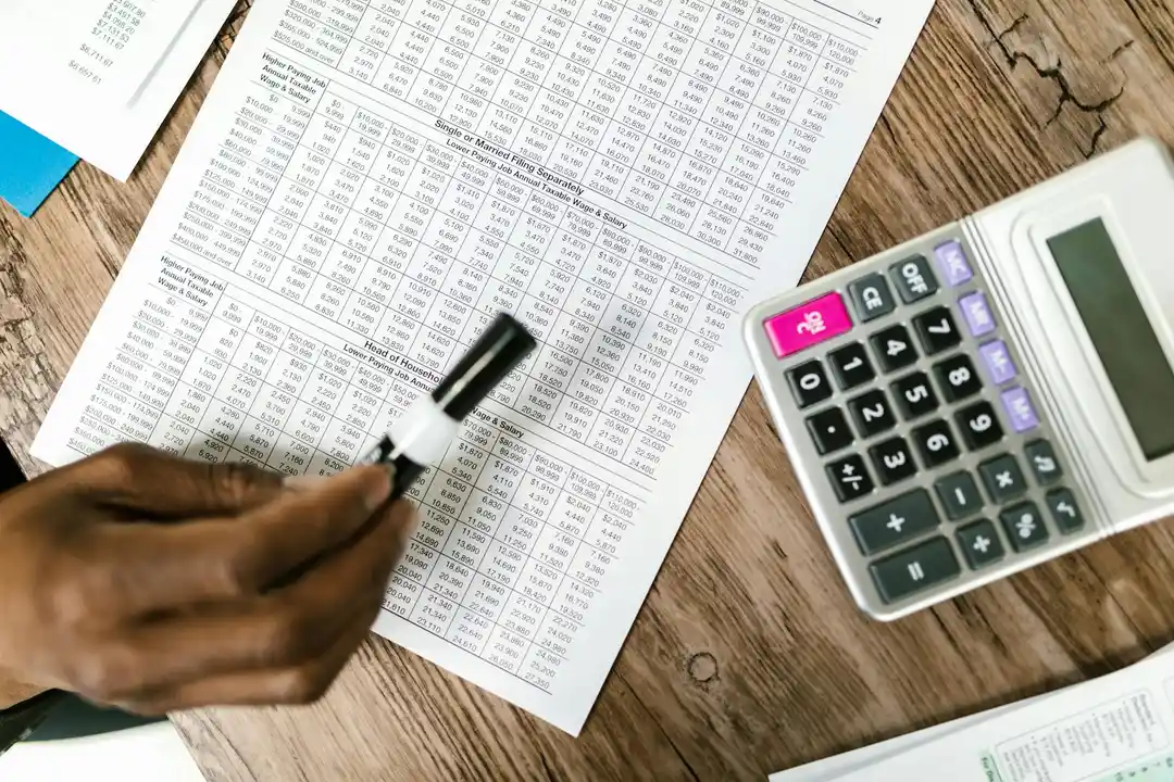 Close-up of a hand using a marker to annotate a printed financial document with dense tables, a calculator nearby on a wooden desk.