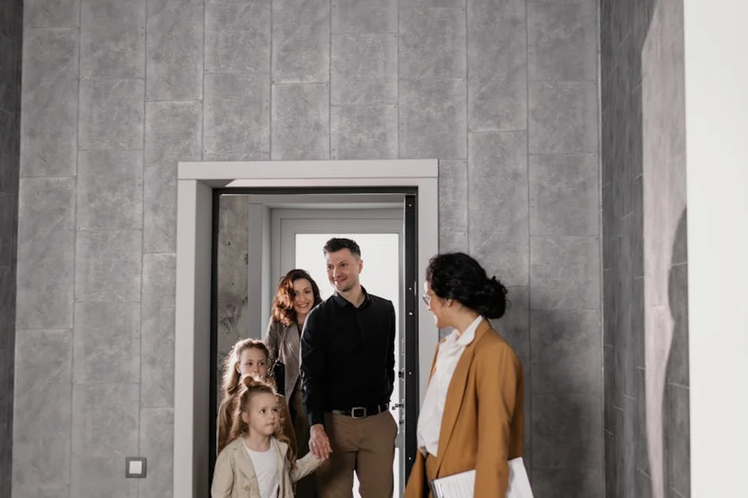 A host in a tan blazer welcomes a family of four at a doorway inside a contemporary building.