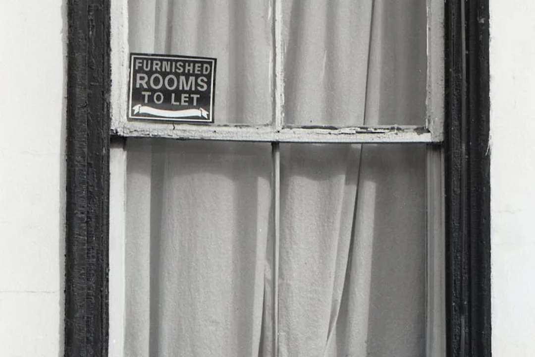 Black-and-white photo of a window with a small sign reading 'FURNISHED ROOMS TO LET' in the upper pane.