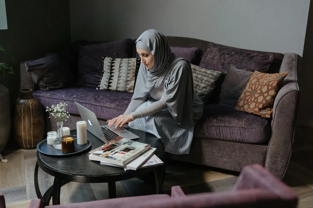 Woman wearing a hijab sitting on a sofa and using a laptop to verify HOA information at home