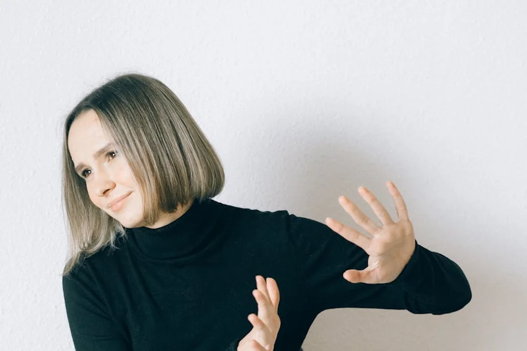 Woman in a black turtleneck raising her hands in a defensive gesture against a plain white wall.