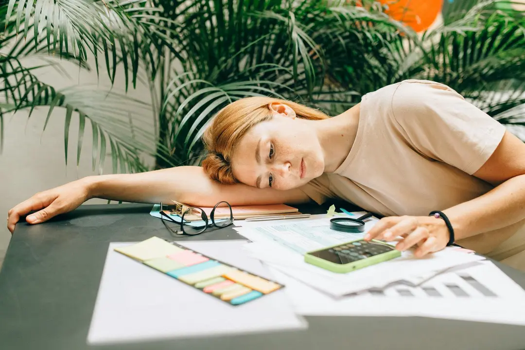 Woman resting her head on a desk, surrounded by paperwork, a calculator, glasses, and color swatches, contemplating HOA fees.