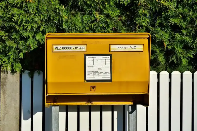 Bright yellow residential mailbox with labels, mounted in front of a white picket fence and dense green hedges.